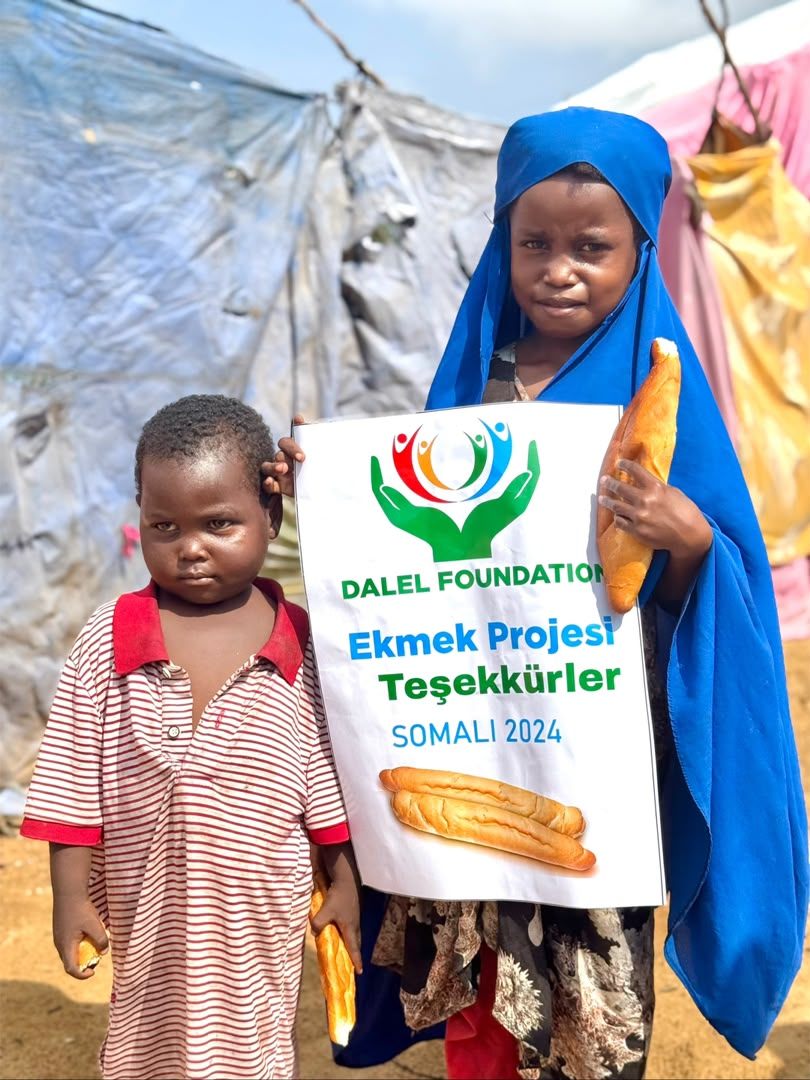 Two children holding fresh bread beside a Dalel Foundation bread project sign in Mogadishu